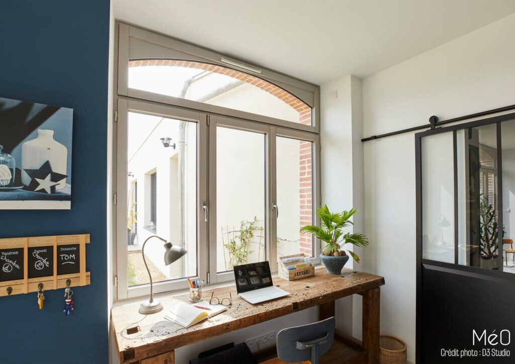 Bureau avec plante et ordinateur près de fenêtre.